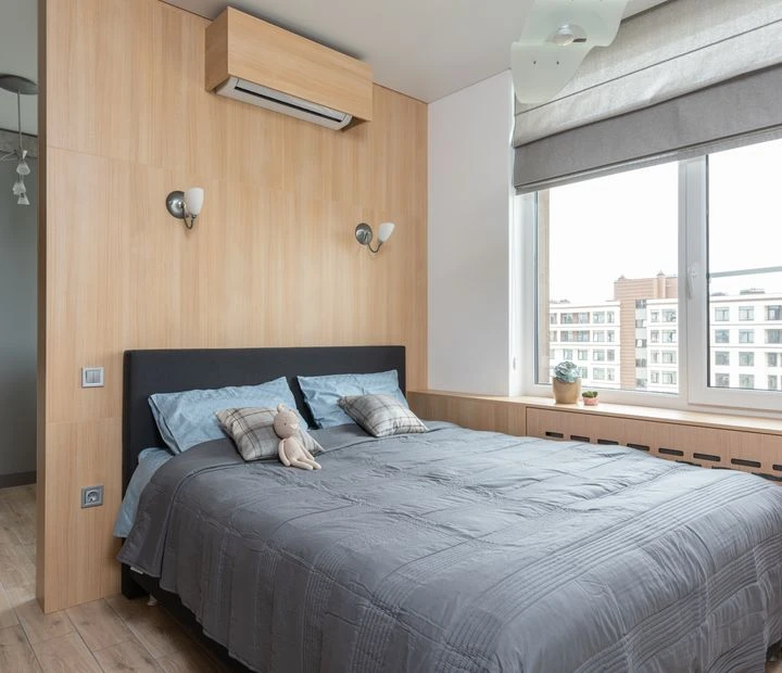 Interior of light contemporary apartment with bed with pillows and soft bunny toy near window with windowsill with decorative objects with jalousie near air conditioner and entrance to kitchen